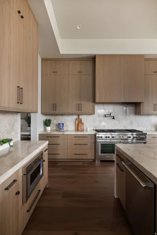Wood-finish cabinetry with matte black hardware, stone countertops and backsplash, stainless steel range with integrated hood, and dark wood-finish flooring