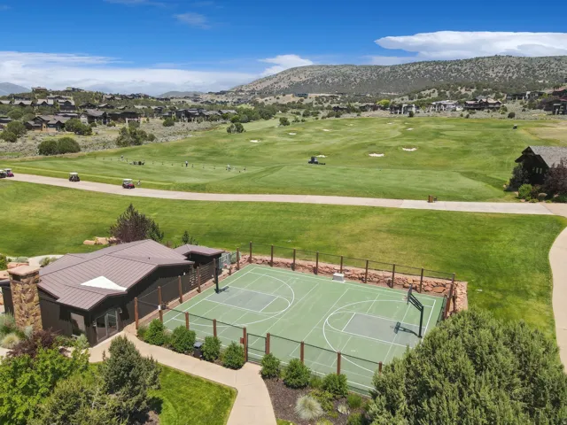 Sports court with perimeter fencing, a detached building featuring stone accents and a metal roof, and a paved pathway, all set within a landscaped area