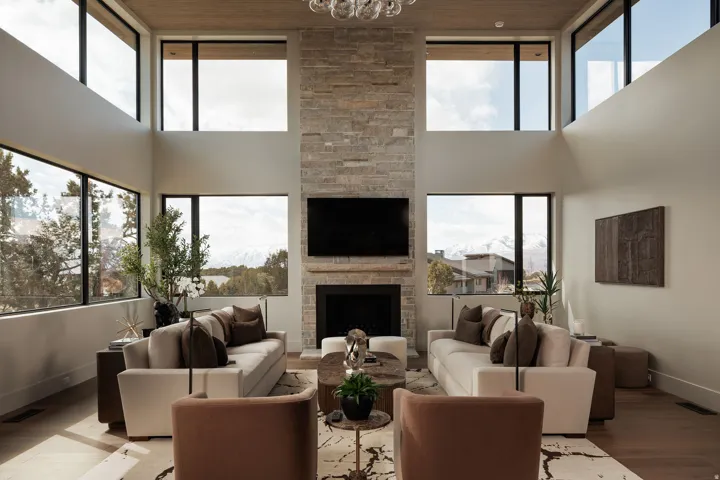 Vaulted ceilings with wood-finish accents, floor-to-ceiling stone fireplace, large black-framed windows, and light wood-finish flooring