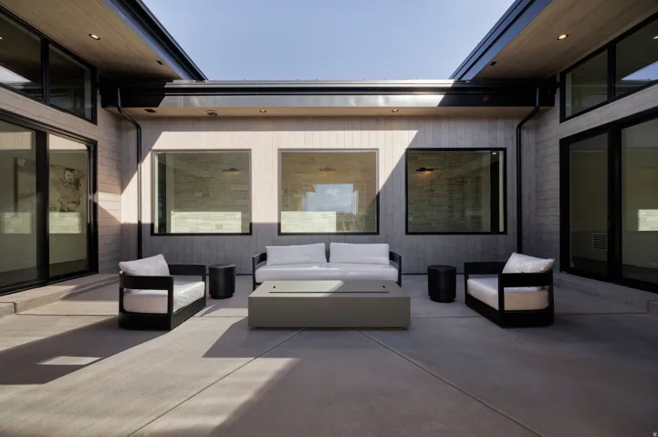 Expansive concrete patio area with a built-in fire feature, framed by walls with large windows and sliding glass doors, and a wood-finish soffit with recessed lighting