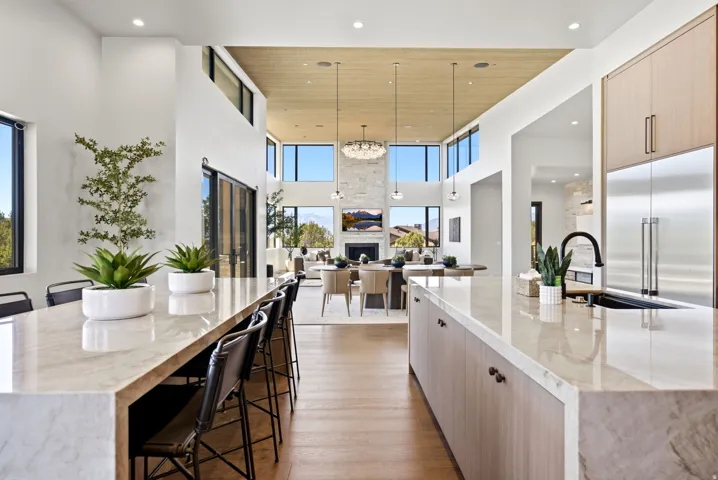Open-concept living space with a double-height ceiling, wood-finish flooring, a large kitchen island with counter seating, and abundant natural light from expansive windows
