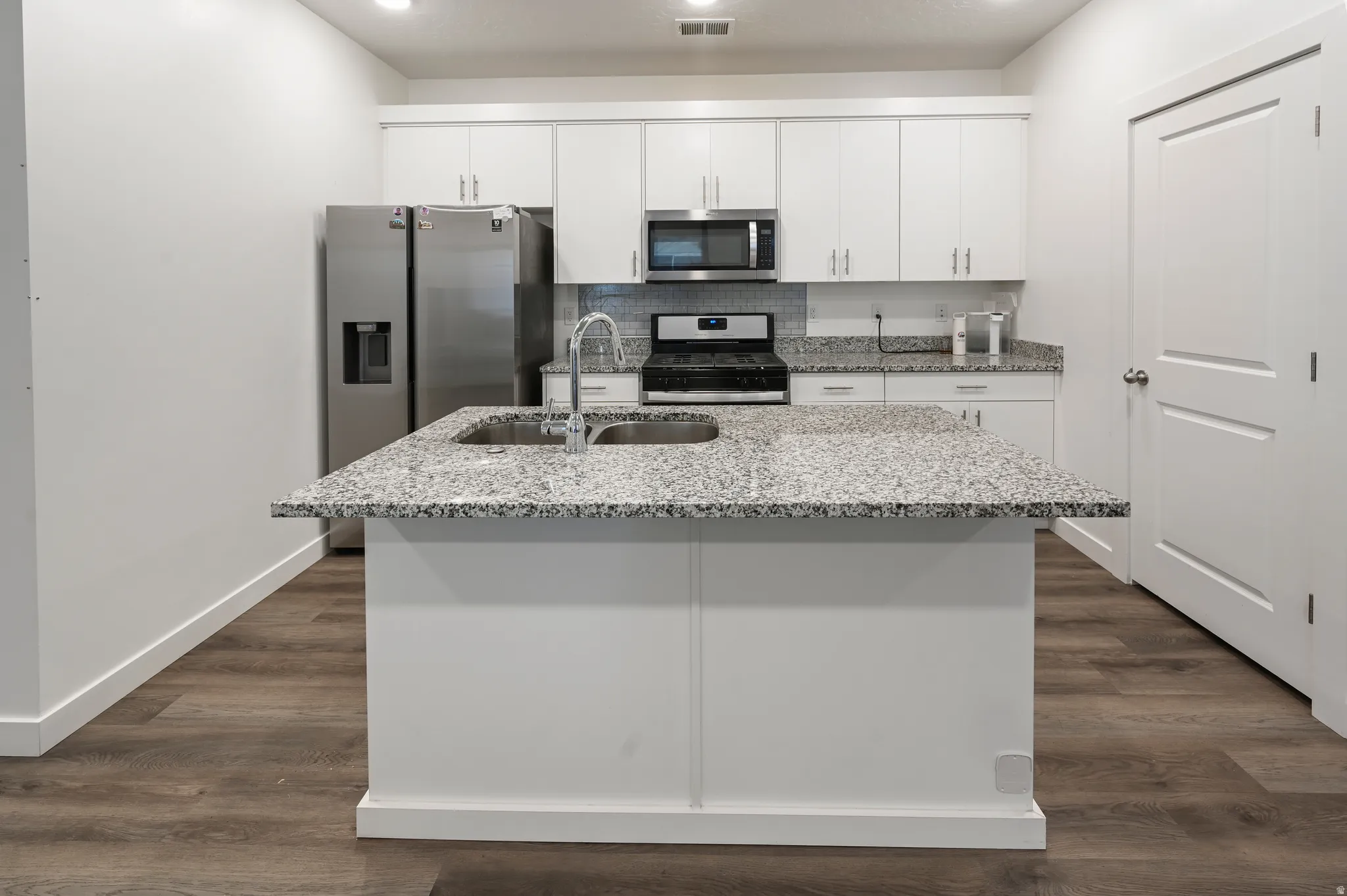 Kitchen featuring light stone countertops, stainless steel appliances, white cabinets, a kitchen island with sink, and dark wood-type flooring