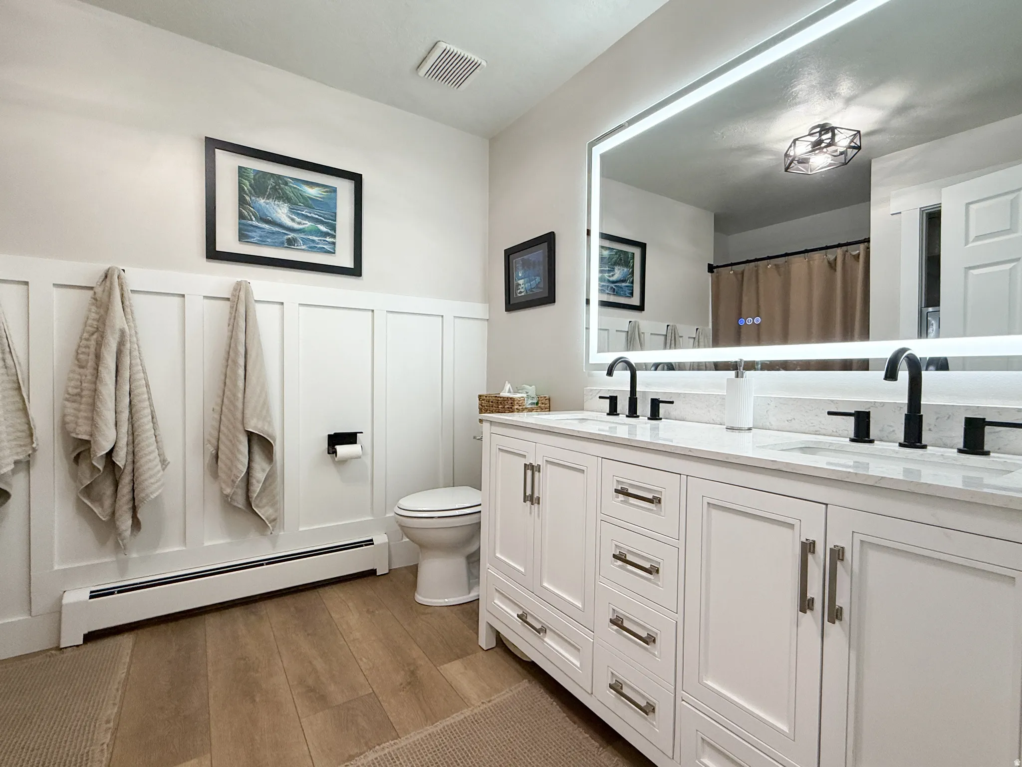 Bathroom with double vanity, a baseboard heating unit, dark wood-type flooring, a wainscoted wall, and a decorative wall