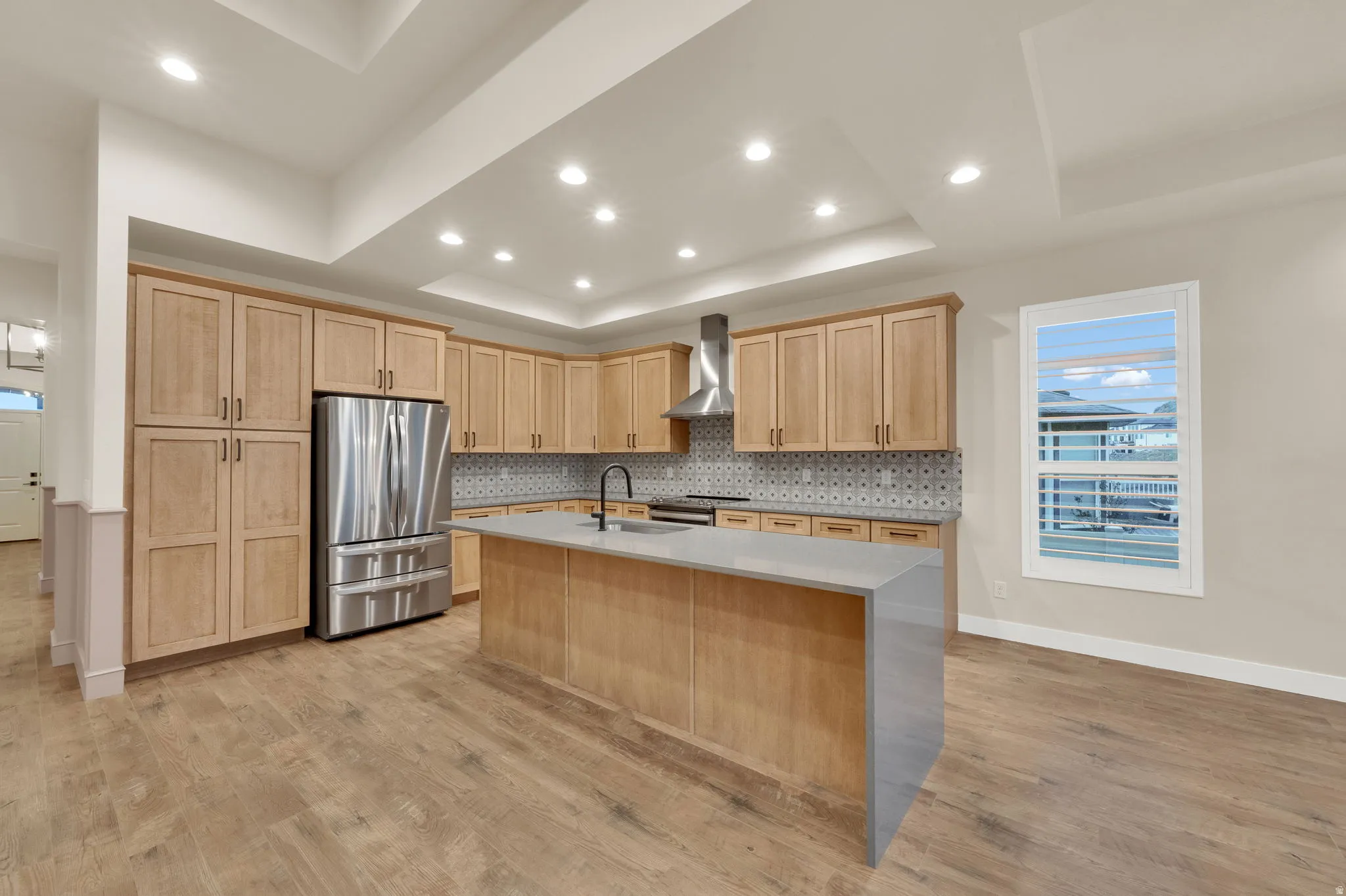 Kitchen with a center island with sink, light stone countertops, stainless steel appliances, light wood-type flooring, and a raised ceiling