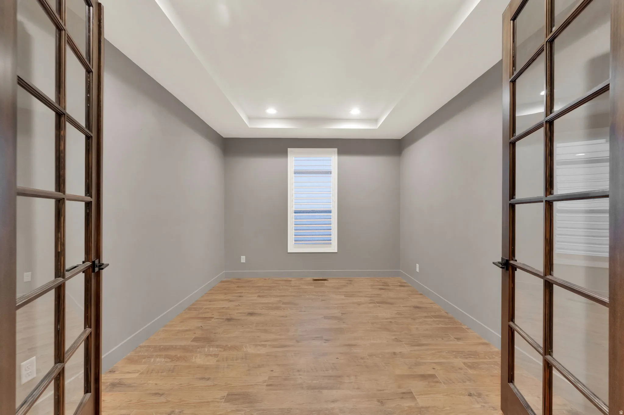 Empty room with french doors, light wood-style floors, a raised ceiling, and recessed lighting