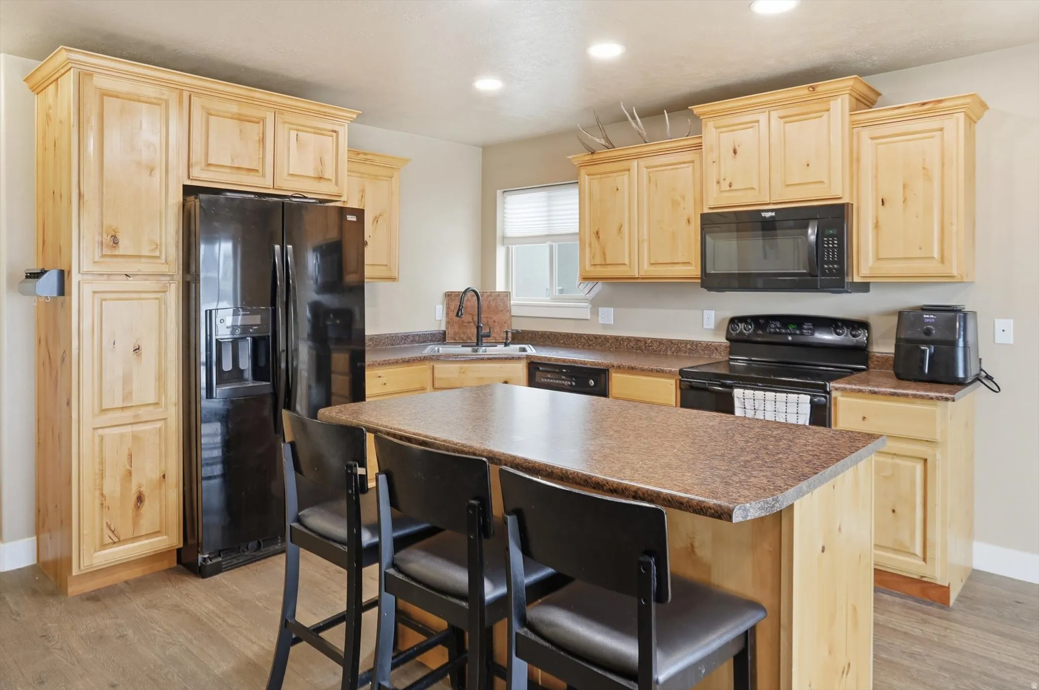Kitchen featuring light wood finish cabinets, black appliances, a center island, dark countertops, and a kitchen bar