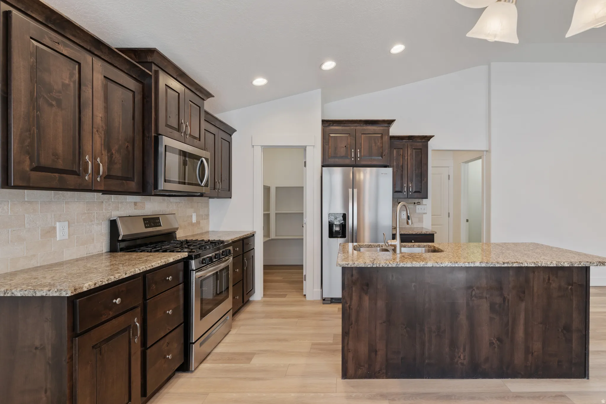 Kitchen featuring dark wood finish cabinetry, stainless steel appliances, lofted ceiling, light wood finished floors, and light stone counters