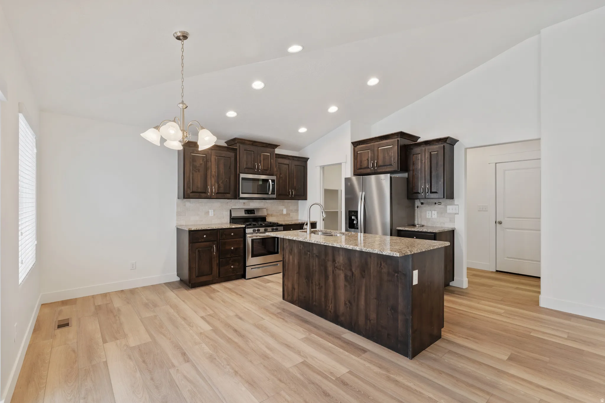 Kitchen featuring dark wood finish cabinetry, vaulted ceiling, decorative backsplash, stainless steel appliances, and light stone counters