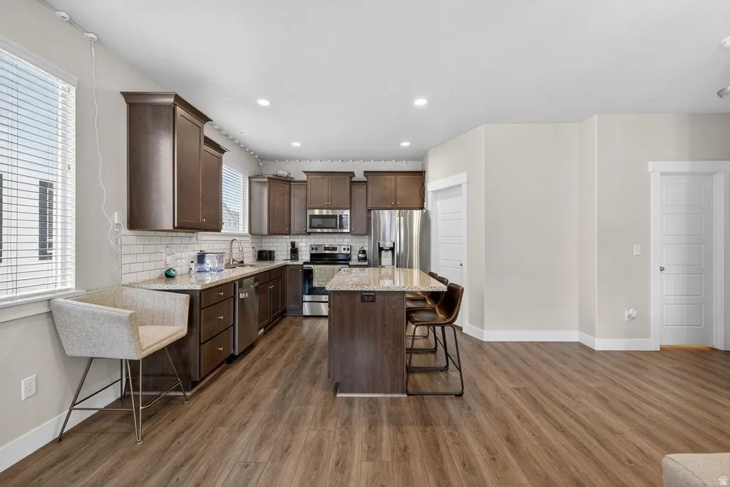 Kitchen featuring dark wood finish cabinets, stainless steel appliances, a kitchen breakfast bar, backsplash, and light stone countertops