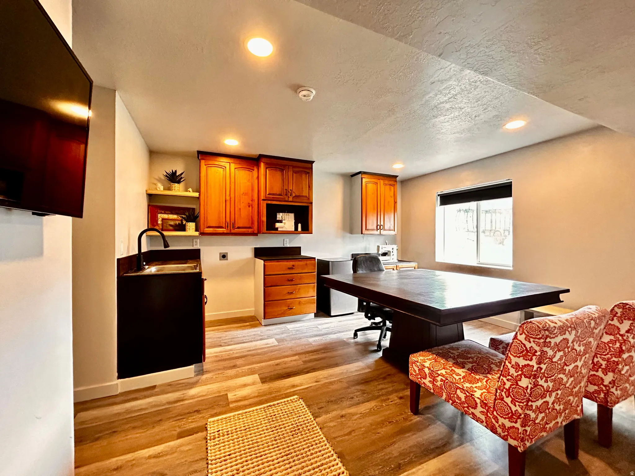 Wood-finish flooring throughout a versatile room featuring a built-in wet bar with a black cabinet and faucet, natural wood cabinetry, recessed lighting, and a window with a black roller shade