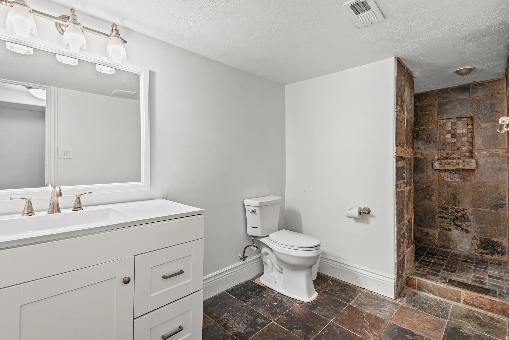 Bathroom with tiled shower, vanity, stone tile flooring, and a textured ceiling