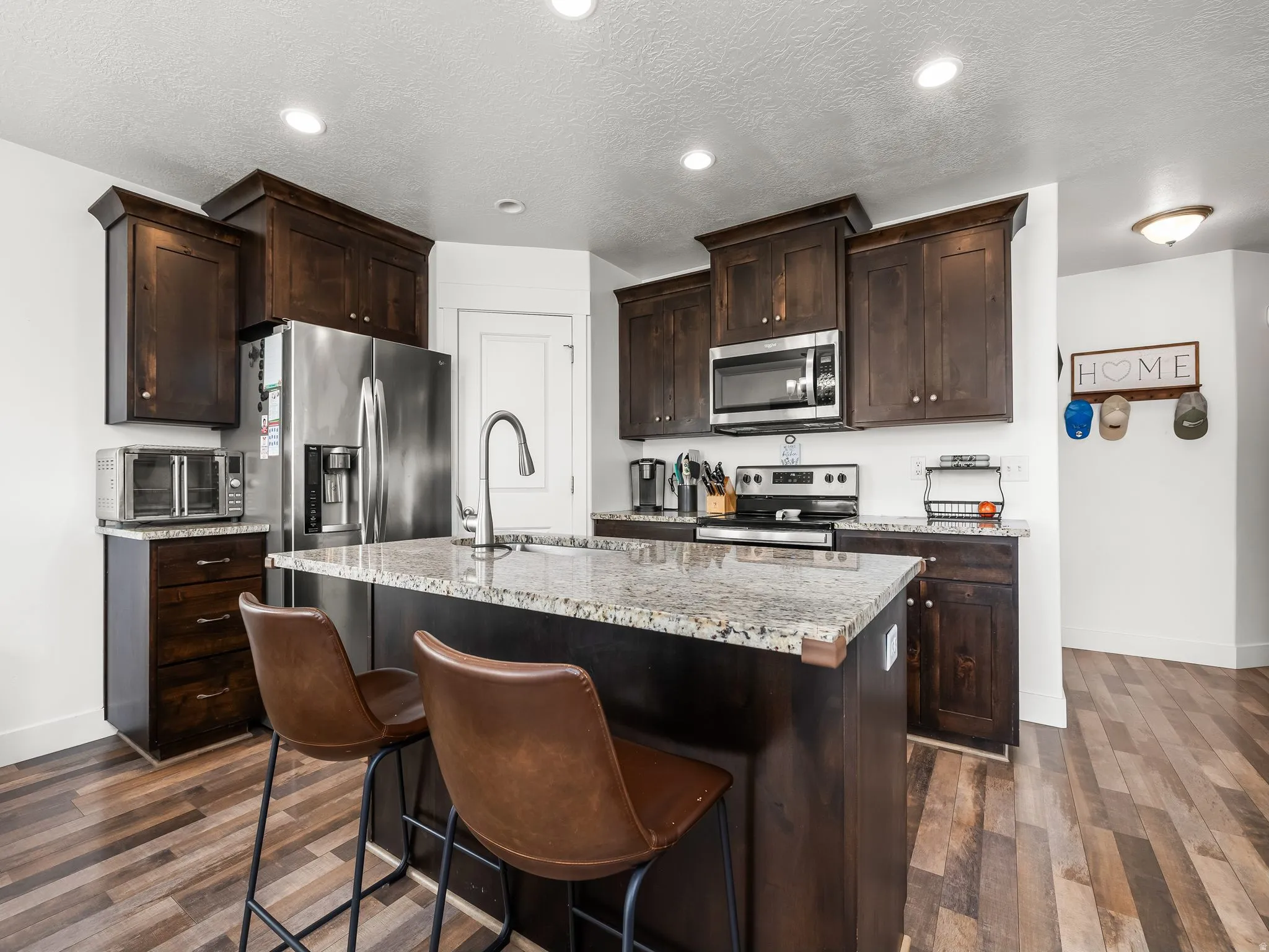 Kitchen featuring dark wood finish cabinets, stainless steel appliances, a center island with sink, dark wood-type flooring, and light stone counters