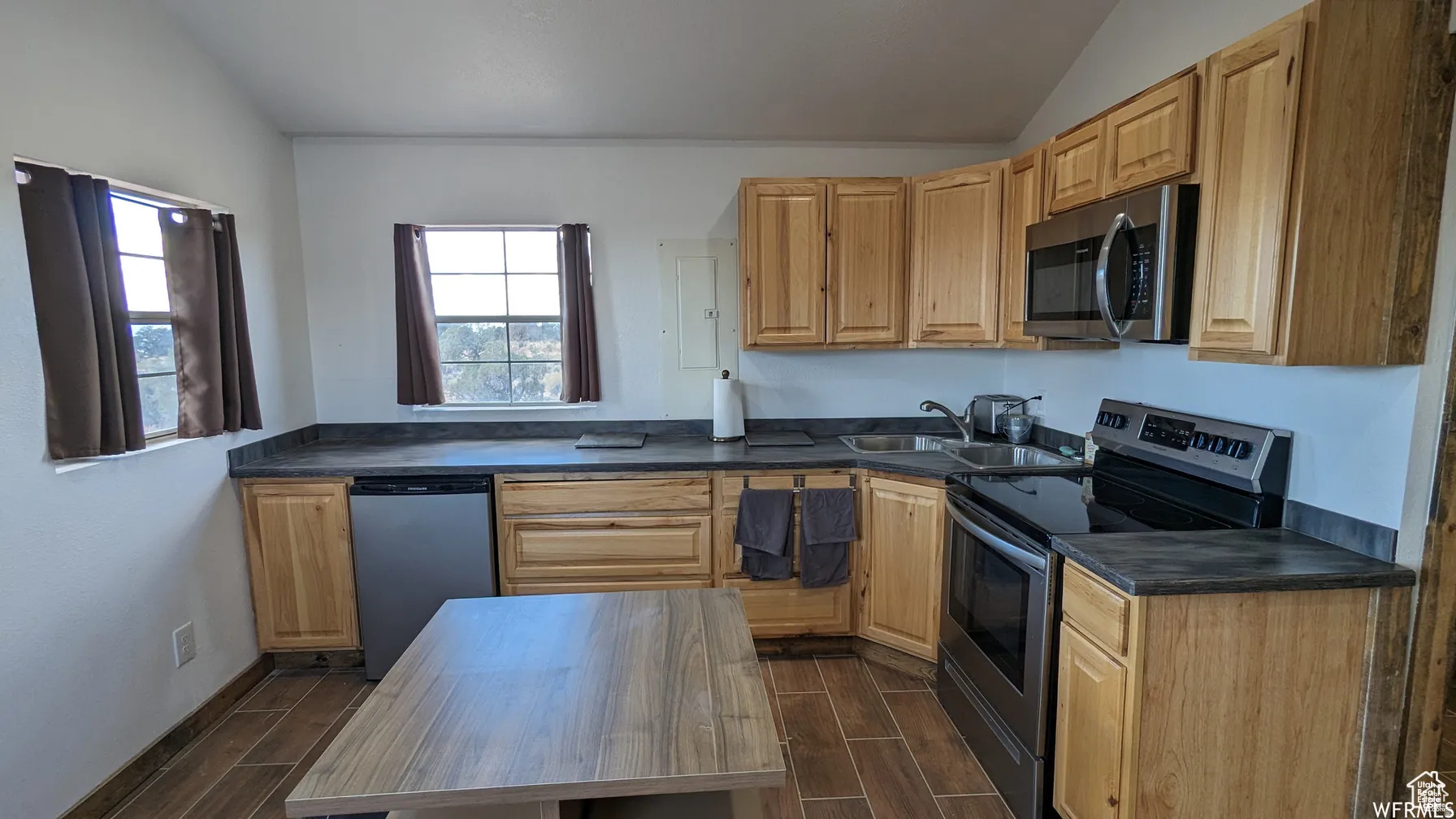 Kitchen featuring stainless steel appliances, vaulted ceiling, dark countertops, wood finish floors, and light wood finish cabinetry
