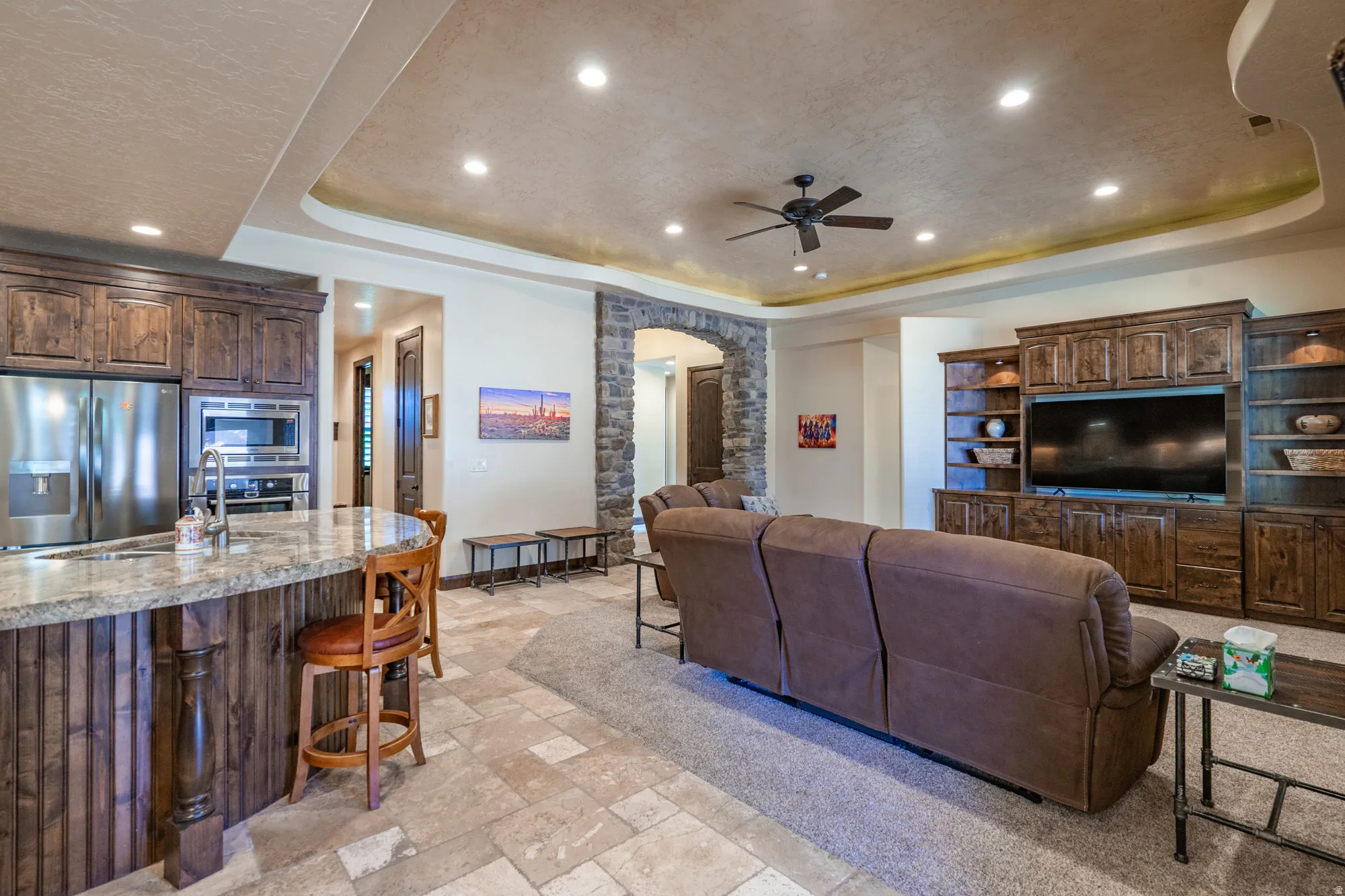 Living room with a raised ceiling, arched walkways, stone tile floors, recessed lighting, and ceiling fan