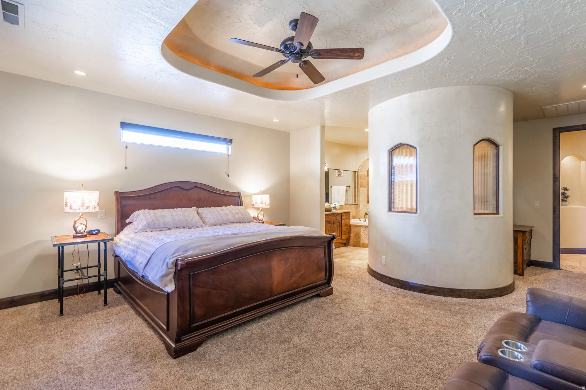 Bedroom with a tray ceiling, light colored carpet, a ceiling fan, connected bathroom, and arched walkways
