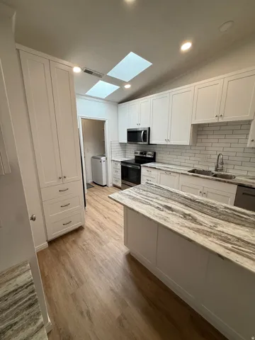 Kitchen featuring vaulted ceilings with skylights, white shaker cabinetry, stainless steel appliances, wood-finish flooring, and a central island with a patterned countertop