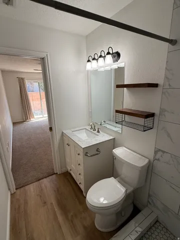 White vanity with a light-toned countertop, mirrored backsplash, wood-finish flooring, and wall-mounted shelving
