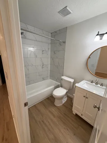 Wood-finish flooring throughout, white vanity with integrated sink, contemporary wall-mounted light fixture, oval mirror, and white subway tile bath surround