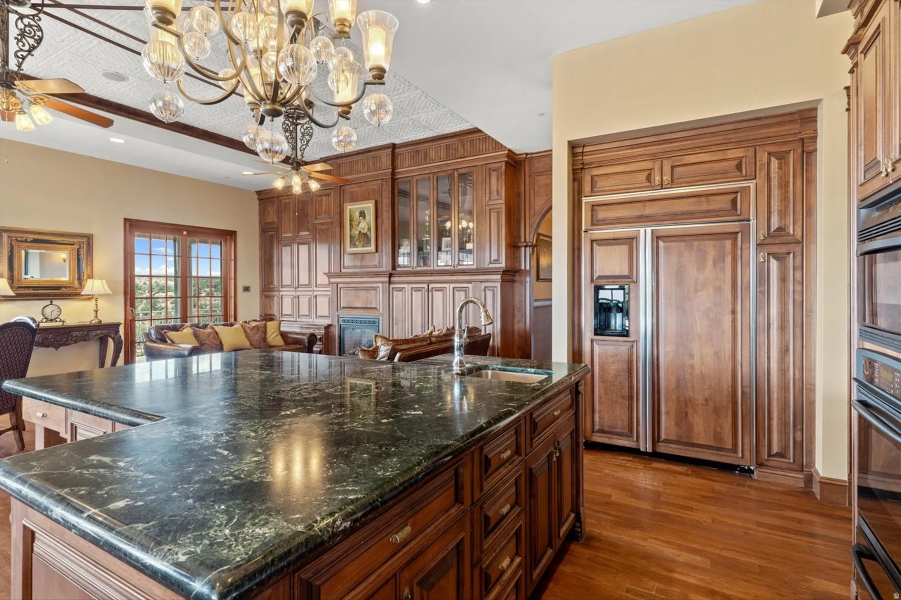 Island with dark stone countertop and integrated sink, wood-paneled cabinetry and appliances, wood-finish flooring, and recessed ceiling with decorative chandeliers