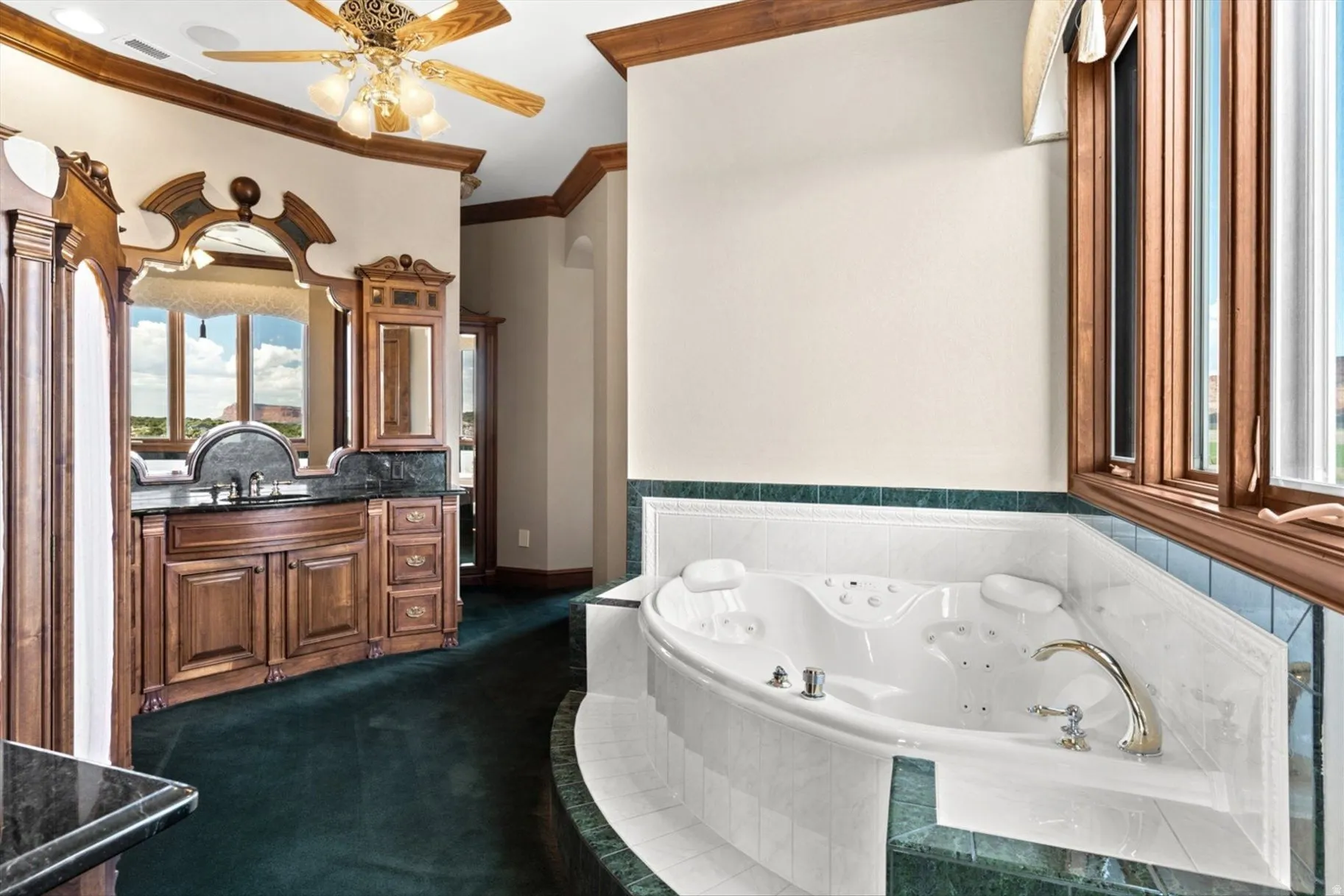 Jetted soaking tub with tile surround, wood-framed windows, double vanity with dark stone countertop, ornate wood cabinetry, and crown molding