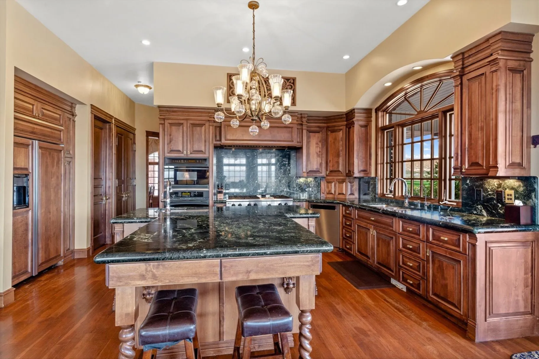 Spacious kitchen featuring rich wood cabinetry, dark stone countertops, wood-finish flooring, a large center island, and an arched window with intricate muntins