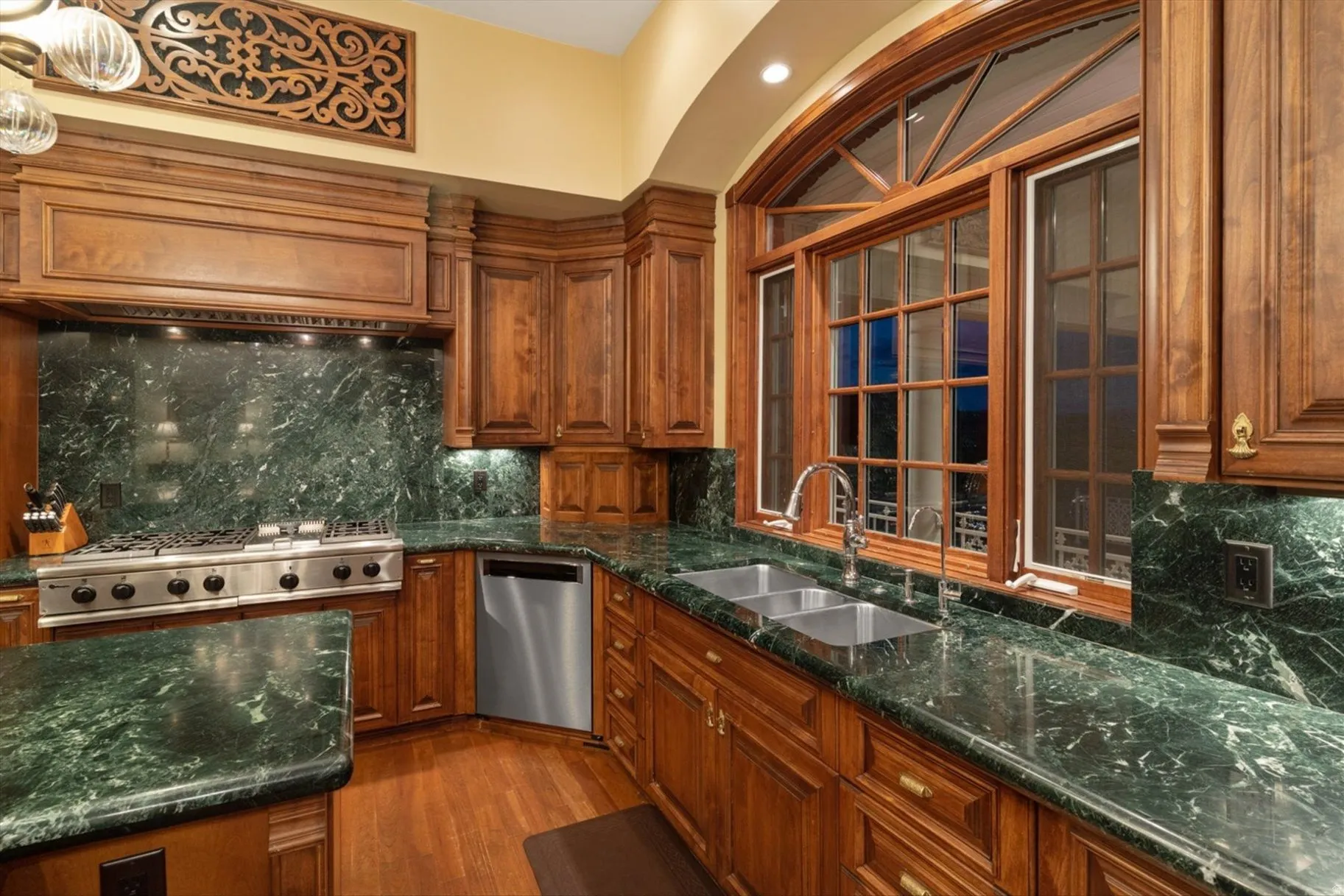 Arched window with wood trim, stainless steel appliances, dual basin sink, wood-finish flooring, and green stone countertops