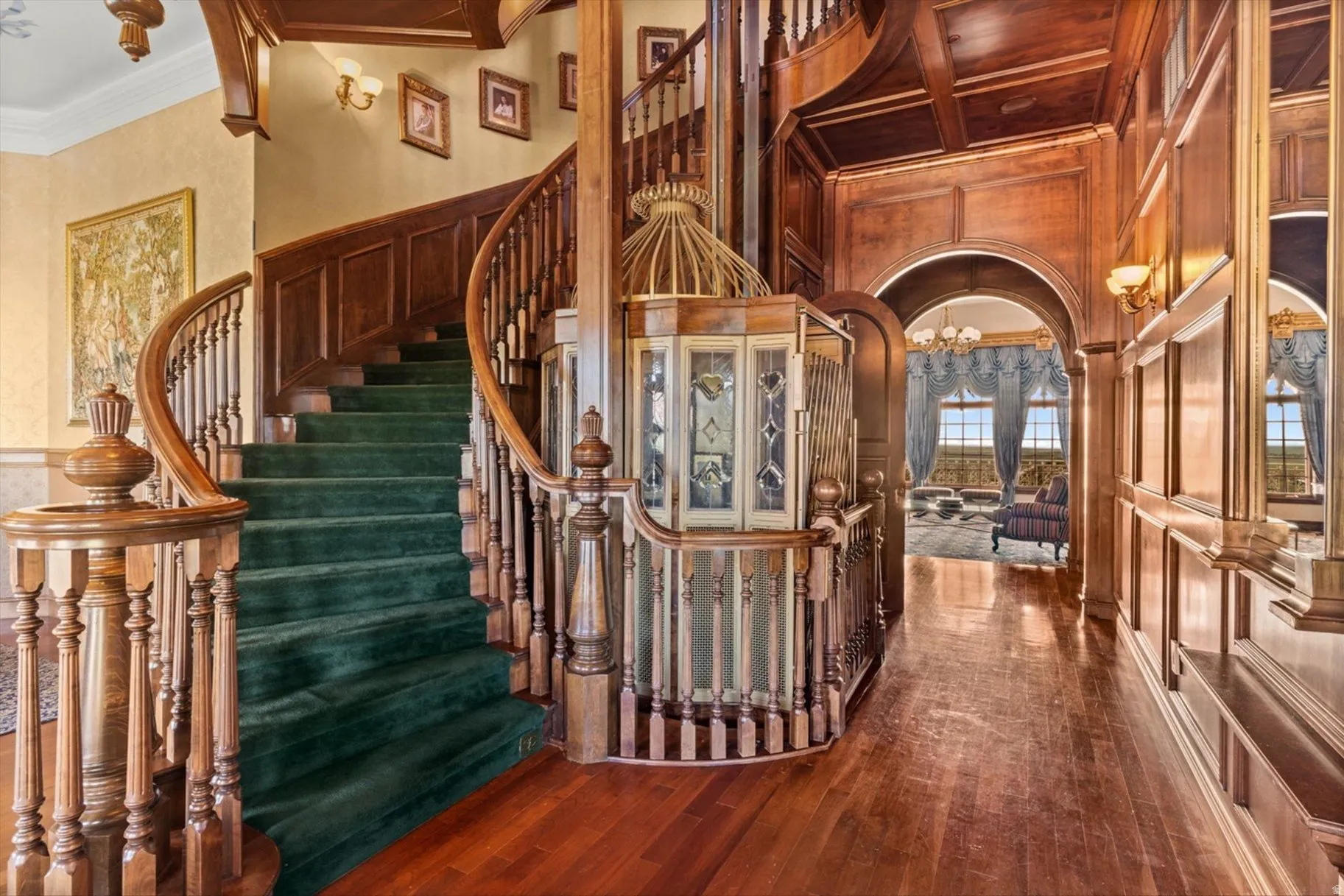 Hardwood flooring, wood paneling, ornate wood staircase with carpeted treads, decorative wall sconces, and an arched entry