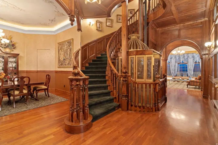 Hardwood flooring throughout, a grand curved staircase with intricate wood detailing, paneled walls, and a decorative enclosed elevator