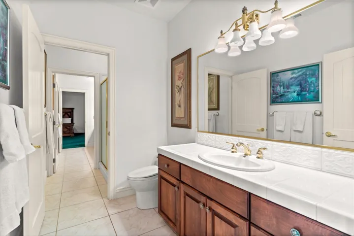 White tile flooring, wood-finish vanity with an integrated sink, polished brass fixtures, and a framed mirror with an overhead light bar