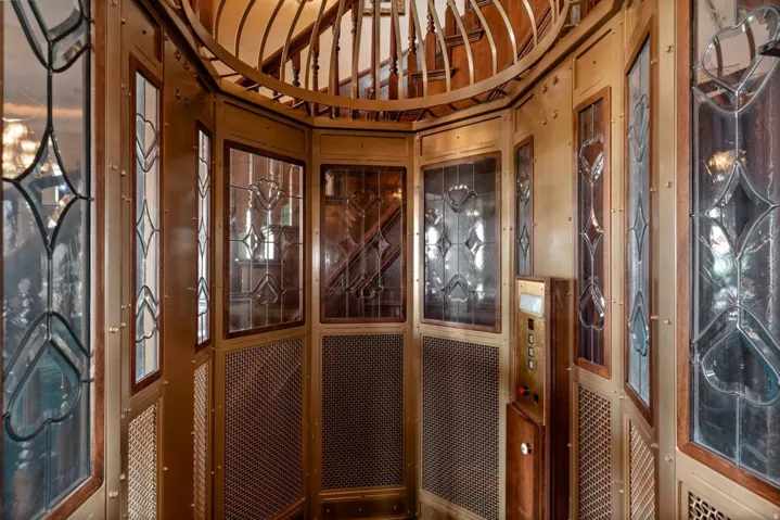 Ornate interior elevator with leaded glass panels, brass-toned metal framing, and a wood-paneled control panel