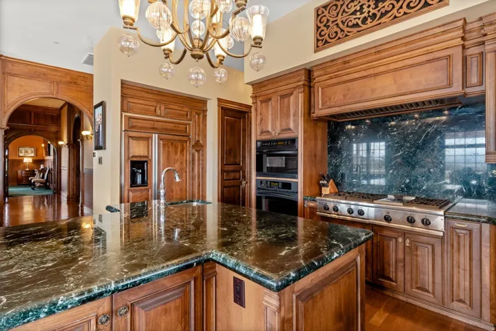 Kitchen featuring wood-finish cabinetry, dark stone countertops, an integrated refrigerator, a stainless steel range with a ventilation hood, and an ornate chandelier