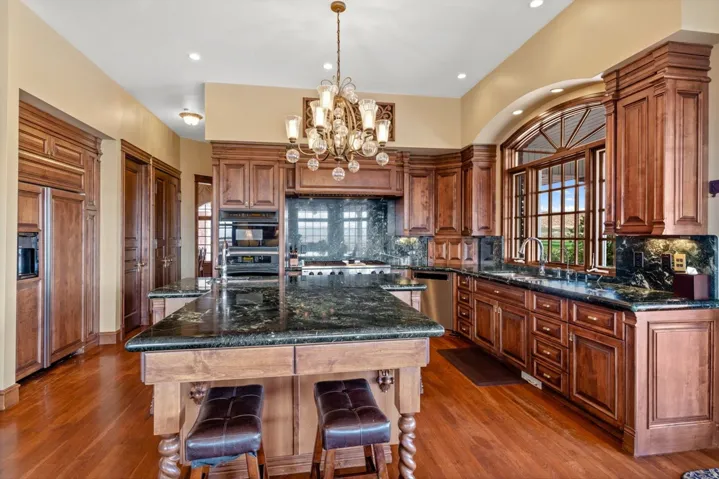 Spacious kitchen featuring rich wood cabinetry, dark stone countertops, wood-finish flooring, a large center island, and an arched window with intricate muntins