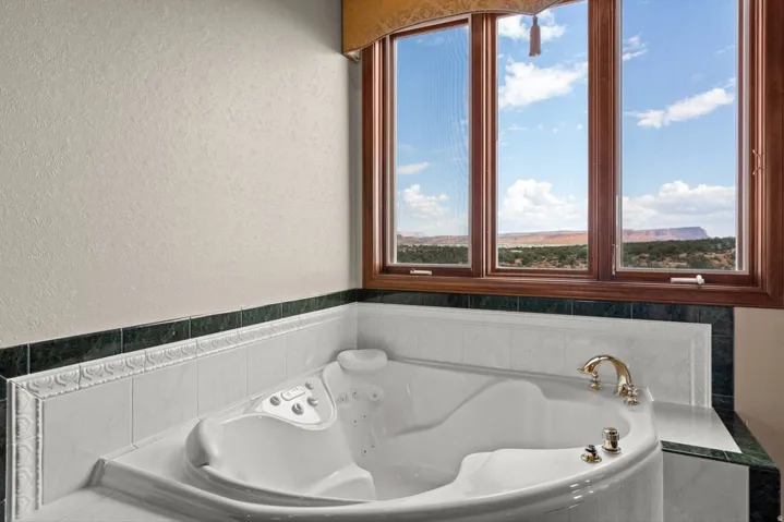 Corner jetted tub with brass fixtures, surrounded by white and green tile, set beneath a large bay window with wood trim