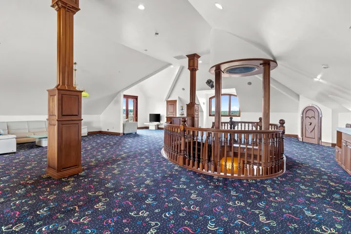 Expansive attic space featuring a large wooden balustrade, multiple windows, a patterned carpet, and built-in cabinetry