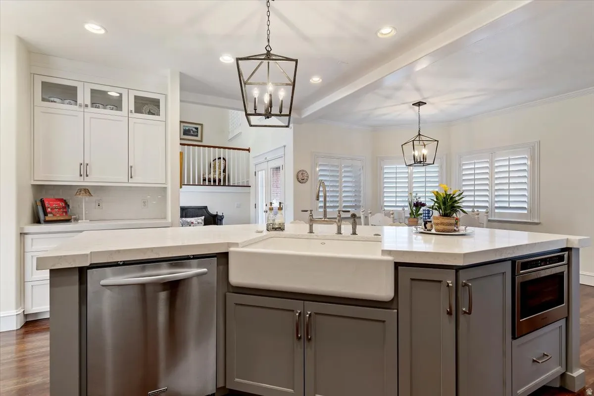 Dual tone kitchen with dark wood finished floors, suspended lighting, stainless steel dishwasher, glass fronted cabinets, and a center island with sink