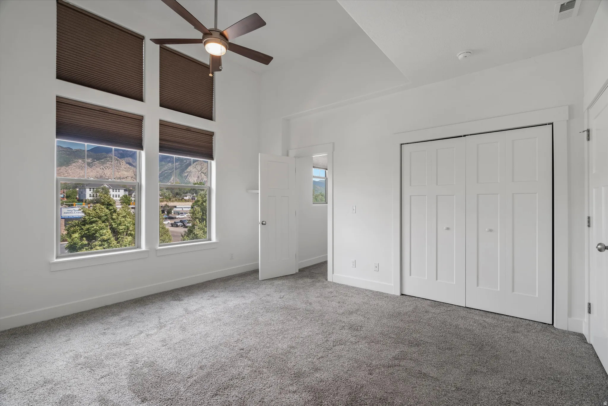 Spacious room featuring multiple large windows with mountain scenery, a ceiling fan, plush gray carpeting, white walls, and a bi-fold closet with panel doors