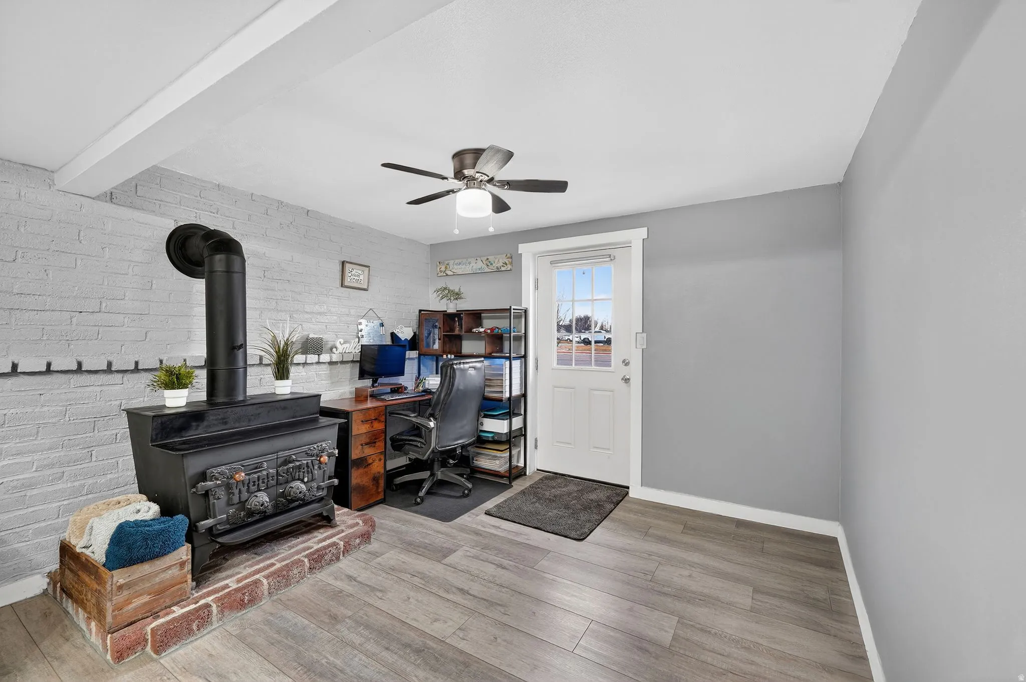 Home office featuring light wood-style floors, brick wall, a wood stove, a ceiling fan, and beam ceiling