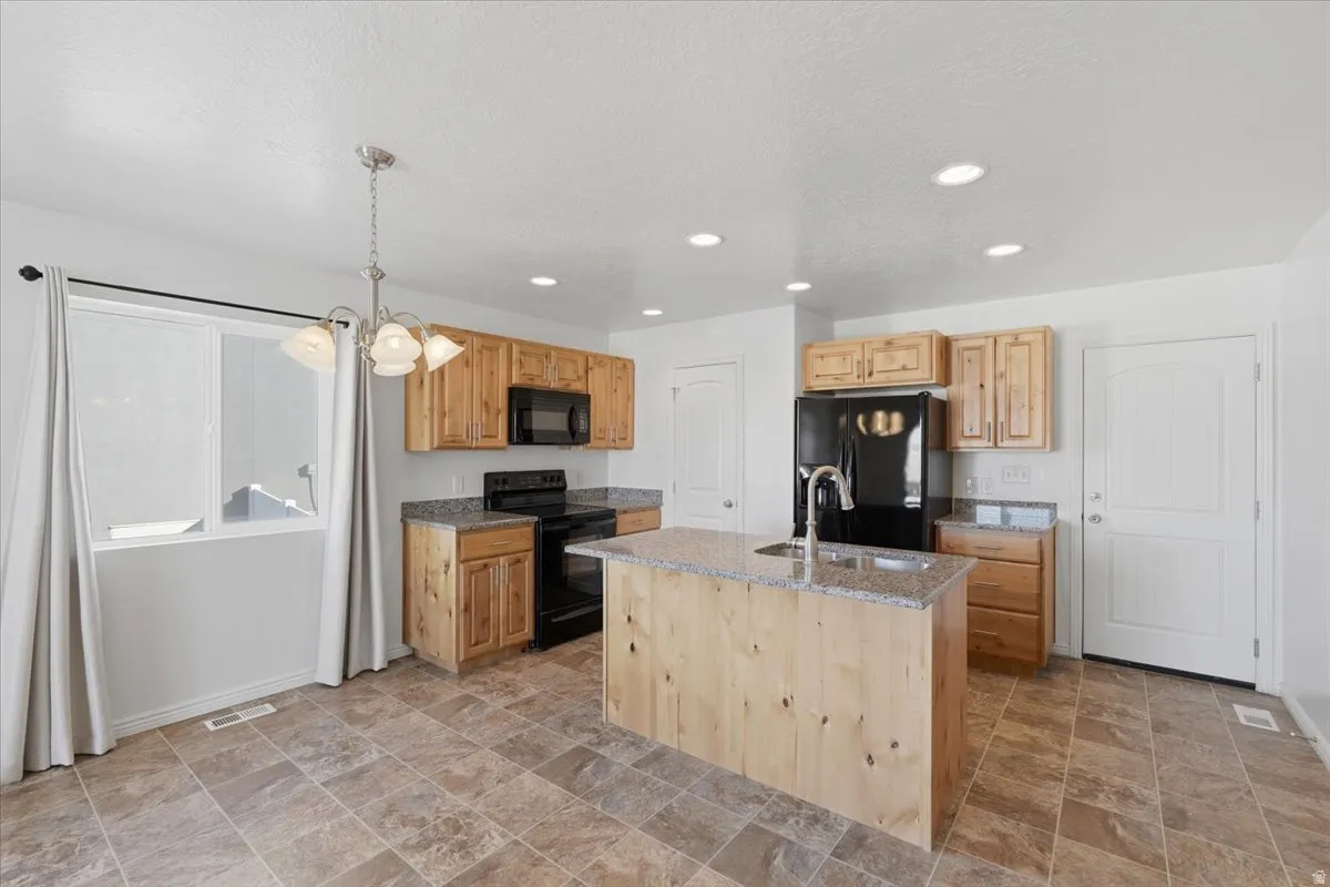 Kitchen with light stone counters, stone finish floors, black appliances, a center island with sink, and light wood finish cabinetry