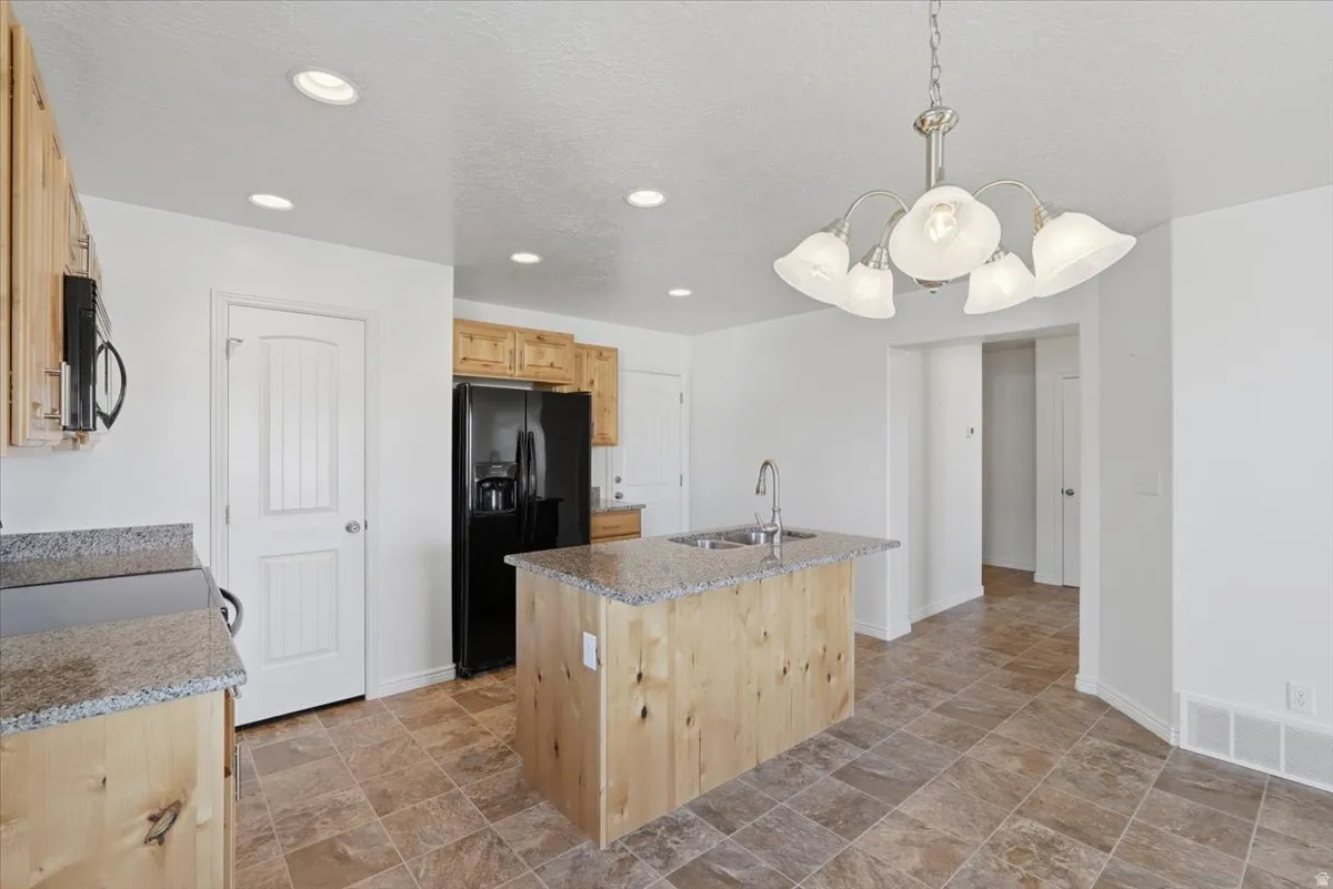 Kitchen with light wood finish cabinetry, stone finish floors, black appliances, an island with sink, and light stone countertops