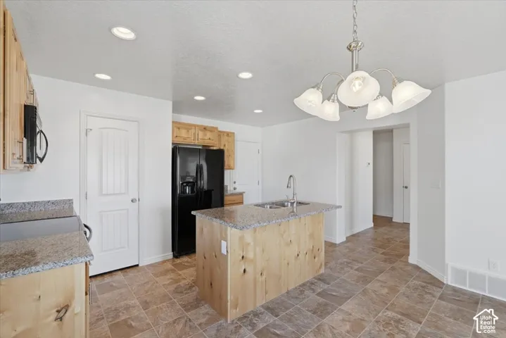 Kitchen with light wood finish cabinetry, stone finish floors, black appliances, an island with sink, and light stone countertops