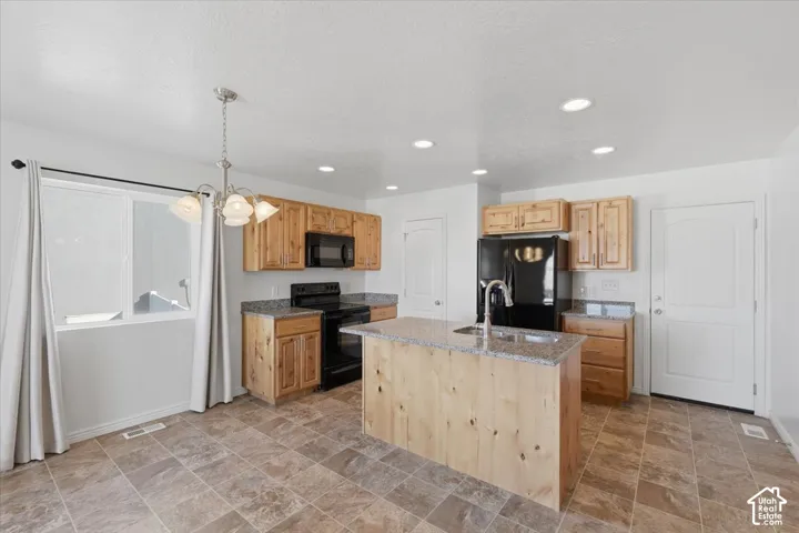 Kitchen with light stone counters, stone finish floors, black appliances, a center island with sink, and light wood finish cabinetry