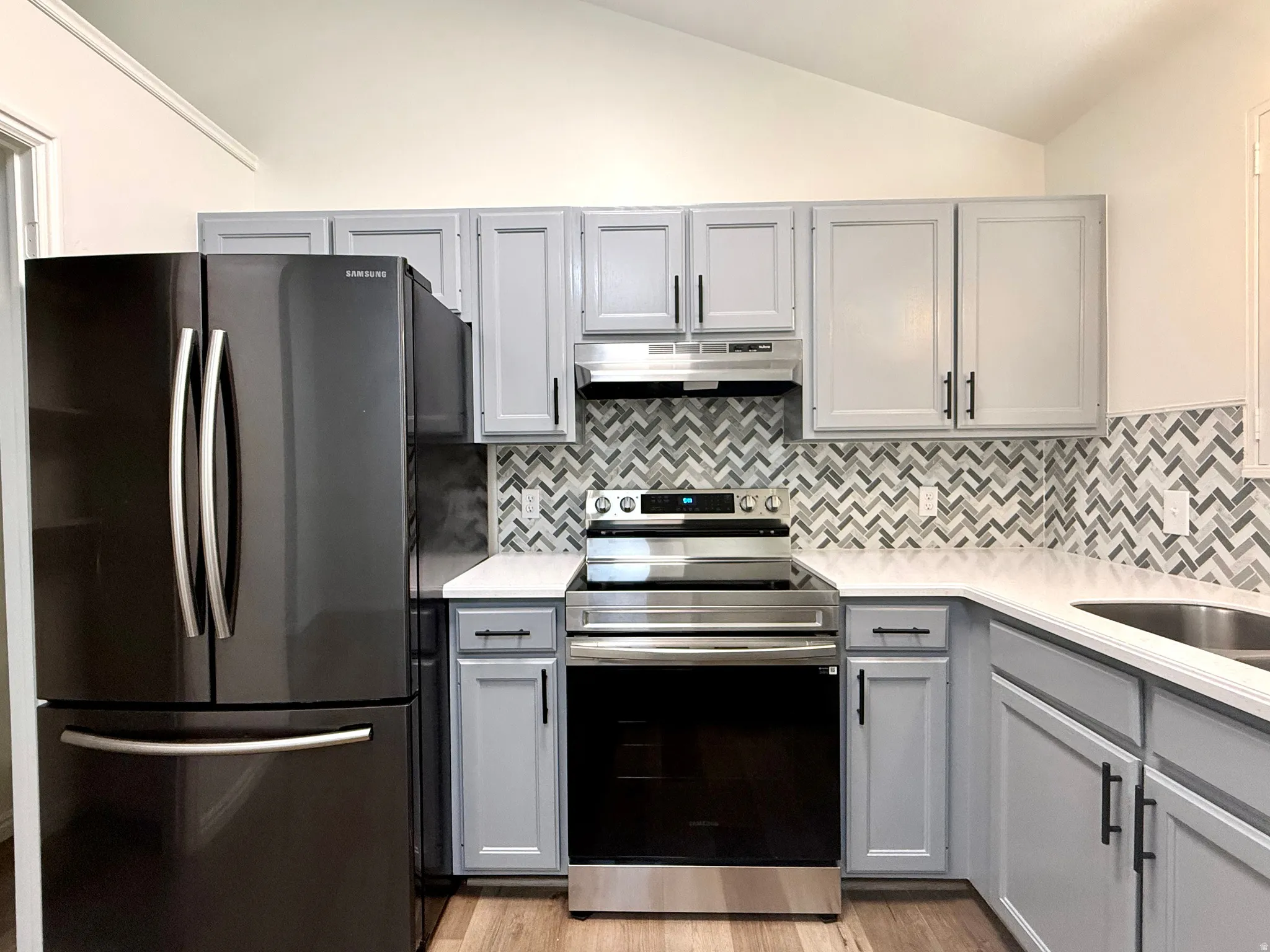 Kitchen with gray cabinetry, freestanding refrigerator, stainless steel electric stove, vaulted ceiling, and light stone counters
