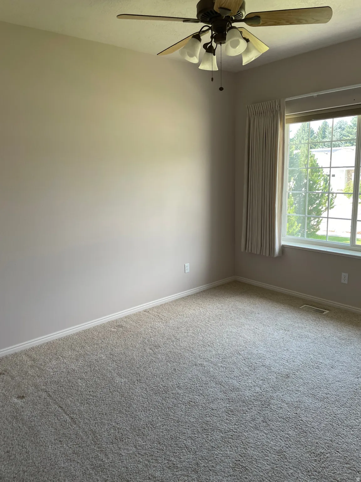Carpeted room with a ceiling fan light fixture, light neutral wall paint, and a large window with window treatments