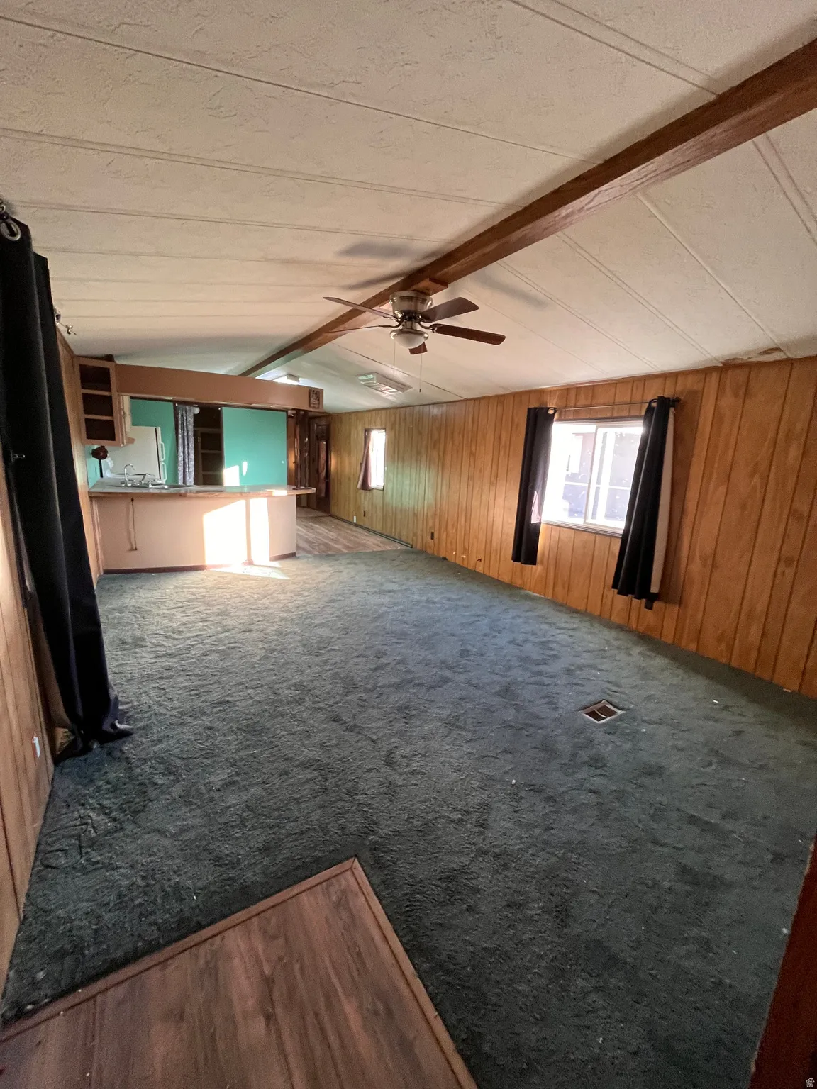 Unfurnished living room with beamed ceiling, wood walls, ceiling fan, and dark colored carpet