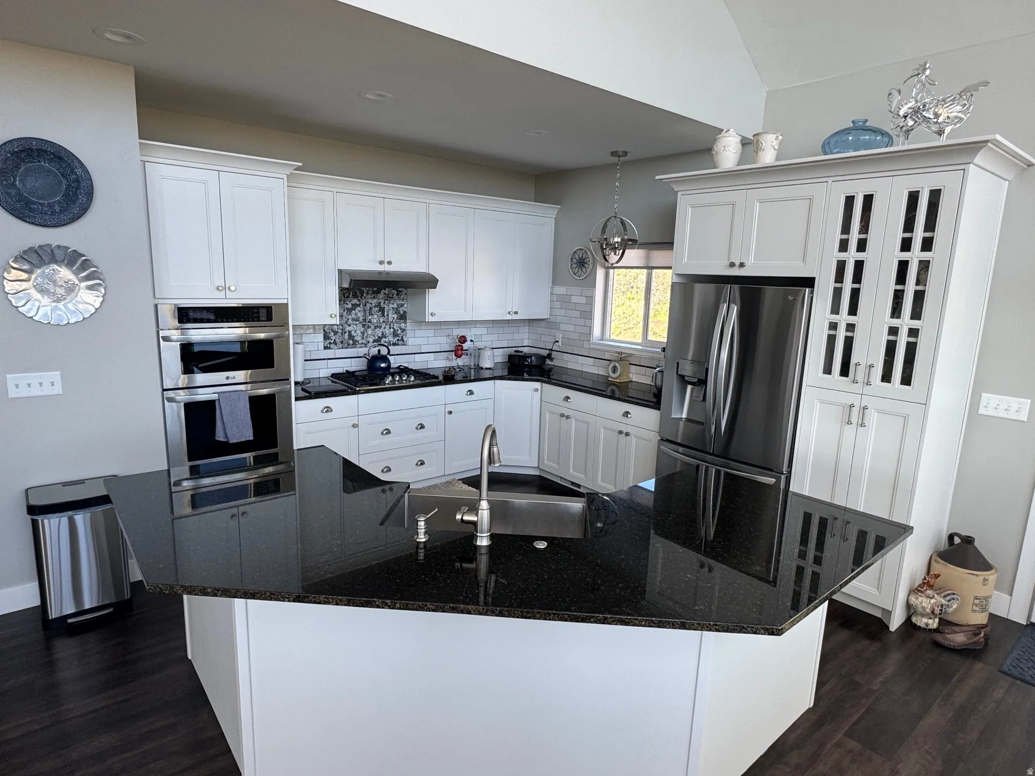 Kitchen featuring an island with a stainless steel undermount sink, black granite countertops, white cabinetry, integrated stainless steel double oven, and a stainless steel gas range with a tile backsplash