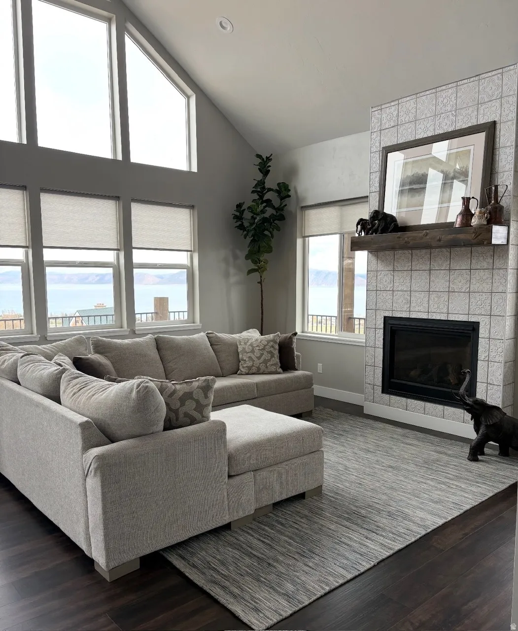 Living area featuring dark wood-finish flooring, a contemporary fireplace with a tiled surround and wood mantle, vaulted ceilings, recessed lighting, and expansive windows showcasing a water backdrop
