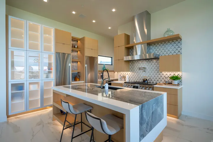 Kitchen with light wood finish cabinets, light stone counters, stainless steel appliances, a kitchen island with sink, and recessed lighting