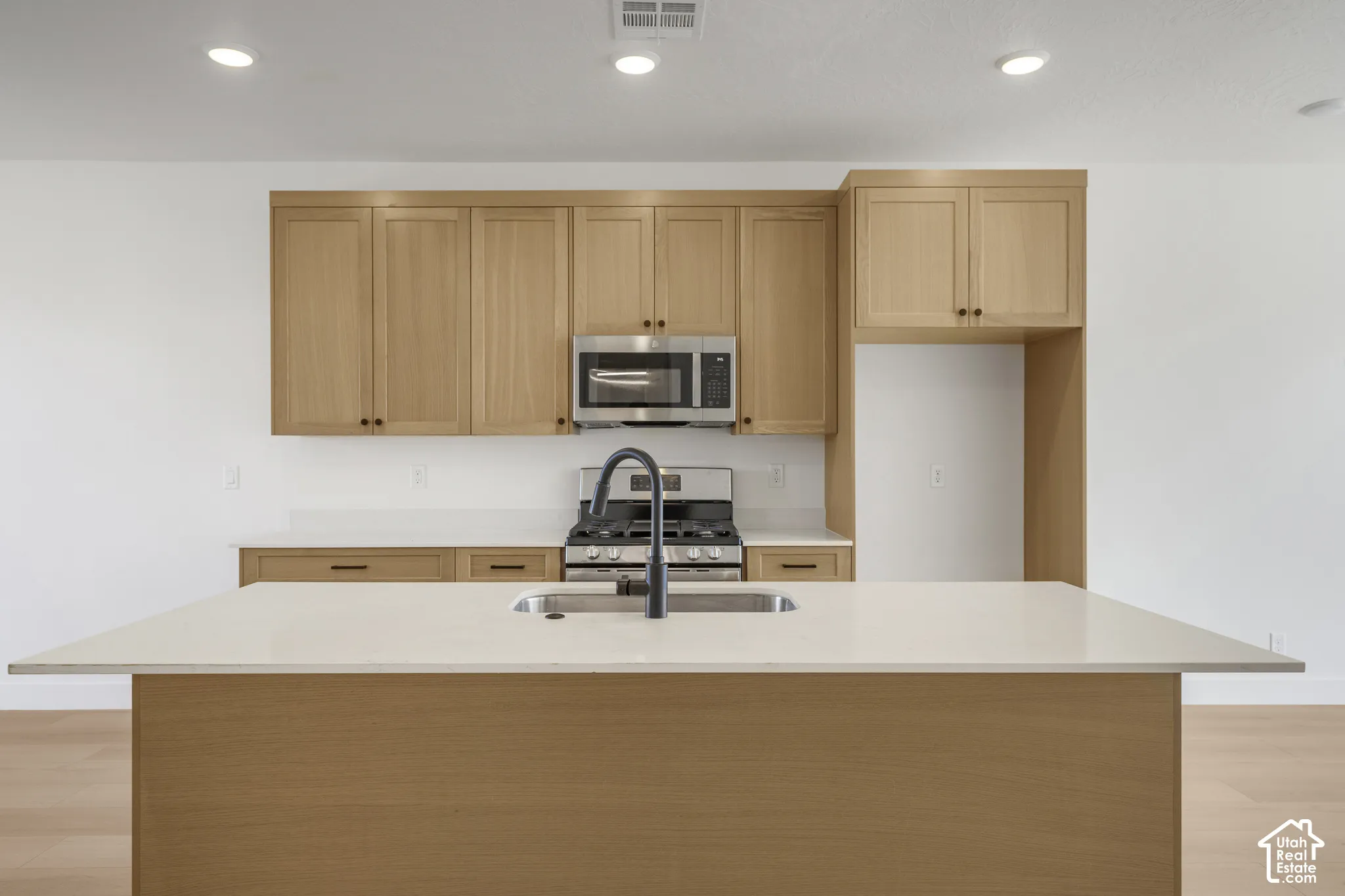 Kitchen featuring light wood-finish cabinetry, white solid-surface countertops, a center island with an undermount sink, a matte black faucet, and stainless steel built-in appliances