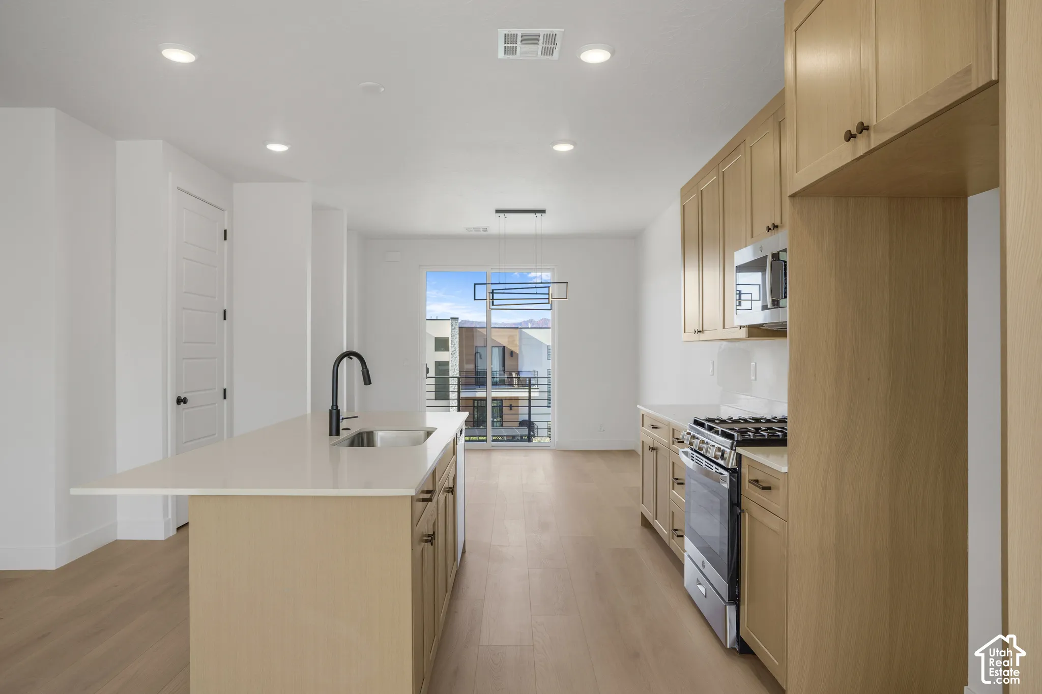 Kitchen featuring light wood-finish cabinetry, stone-surface countertops, a center island with an undermount sink and matte black faucet, integrated stainless steel appliances, and wood-finish flooring