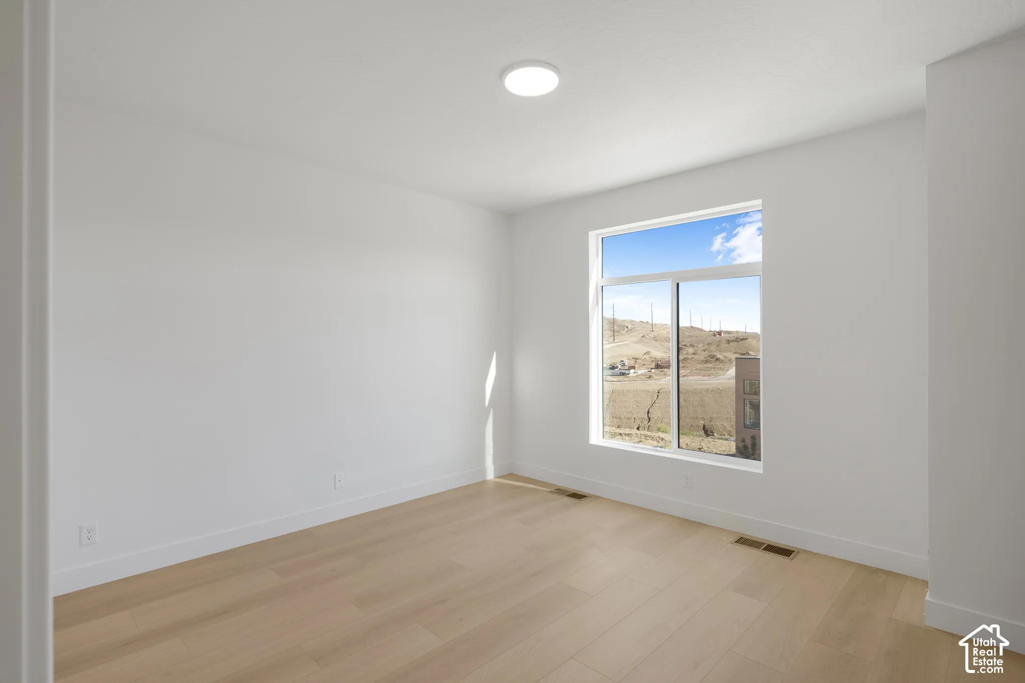 Neutral-toned room featuring light wood-finish flooring, white walls, flush-mount ceiling lighting, and a window with exterior terrain scenery